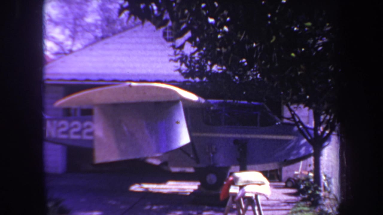 Small airplane under a tarp beside a house and trees.