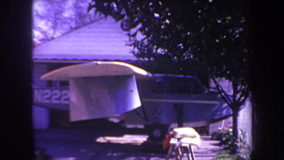 A small single-engine aircraft sits in a residential driveway in the 1969 Yucaipa home-movie footage, with its wing covered by a white tarp and the house roof behind it.