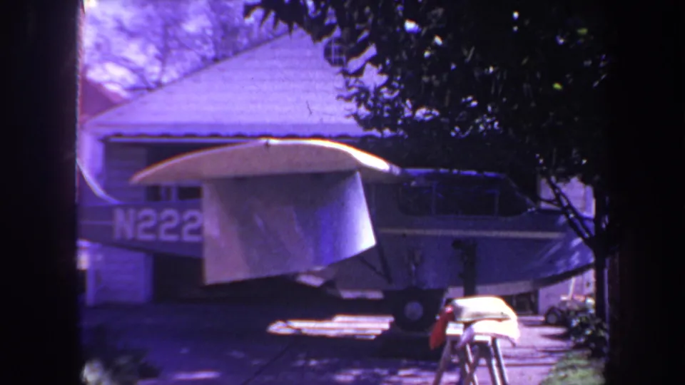 The 8mm frame shows the aircraft tail with partial registration, a white tarp draped over the wing, driveway pavement, and surrounding shrubs in a suburban Yucaipa setting.
