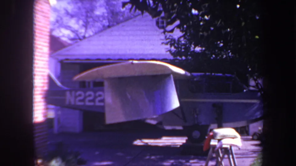 A purple-cast 1969 8mm frame shows the small aircraft parked close to a house, with the wing tarp, partial registration, trees, and vertical foreground framing still visible.