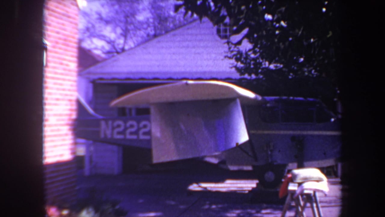 1969 8mm frame from the Yucaipa-area home movie showing a small private airplane parked in a residential driveway, partially covered by a tarp and seen through a dark foreground frame.