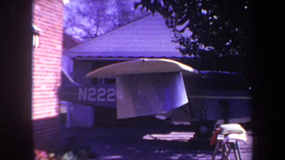 The small aircraft remains parked in the driveway near a brick wall, with partial registration markings visible and a tarp draped over the wing in the 1969 home-movie frame.