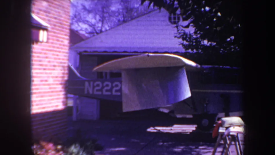 This 1969 Yucaipa-area frame shows the airplane from a similar angle, with the tarp-covered wing, brick exterior wall, driveway surface, and shaded trees visible around it.