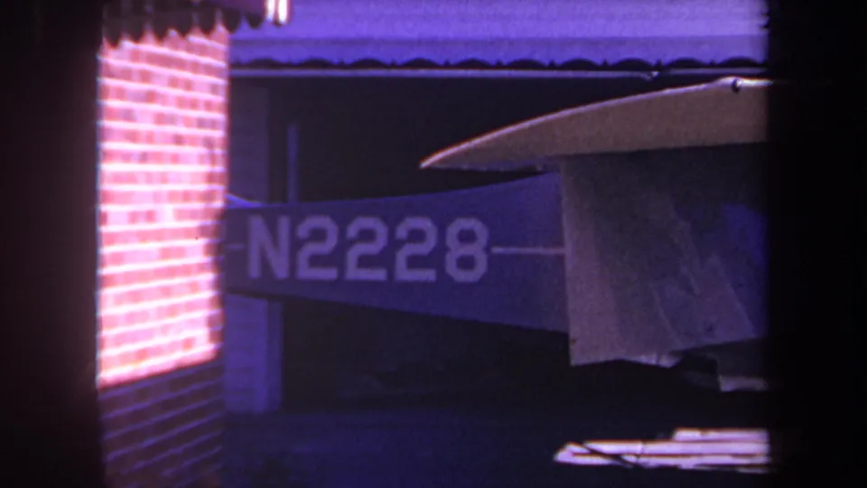 A closer 1969 8mm frame shows the aircraft registration marking more prominently, with the tarp-covered wing, brick wall, driveway shadows, and dark framing still visible.