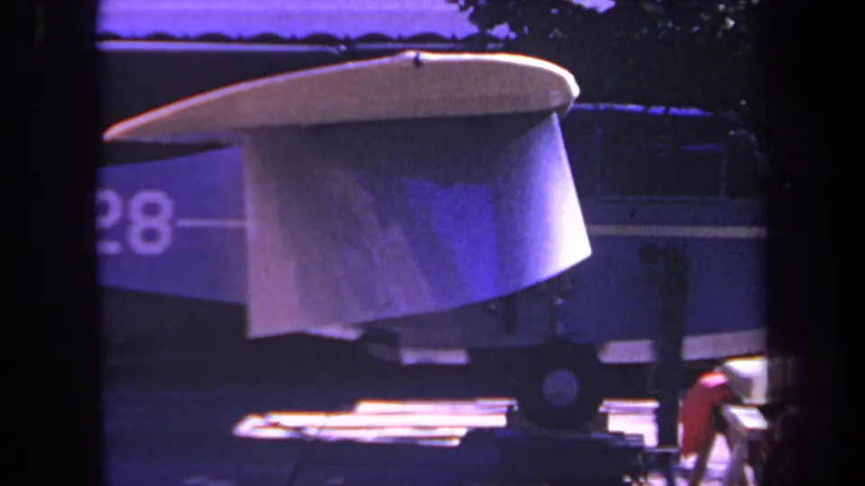 1969 Yucaipa, California 8mm frame showing the aircraft tail from a closer cropped angle, with partial registration, rear wheel, building background, and heavy purple color shift.
