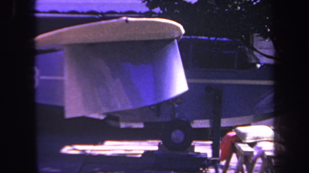 1969 8mm home-movie frame from Yucaipa, California, showing a small blue airplane or aircraft body on blocks or a trailer in a residential driveway, with dark window-like framing in front.