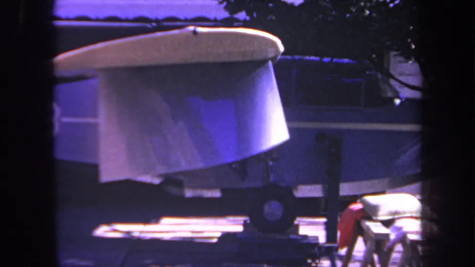 1969 8mm home-movie frame from Yucaipa, California, showing a small blue airplane or aircraft body on blocks or a trailer in a residential driveway, with dark window-like framing in front.