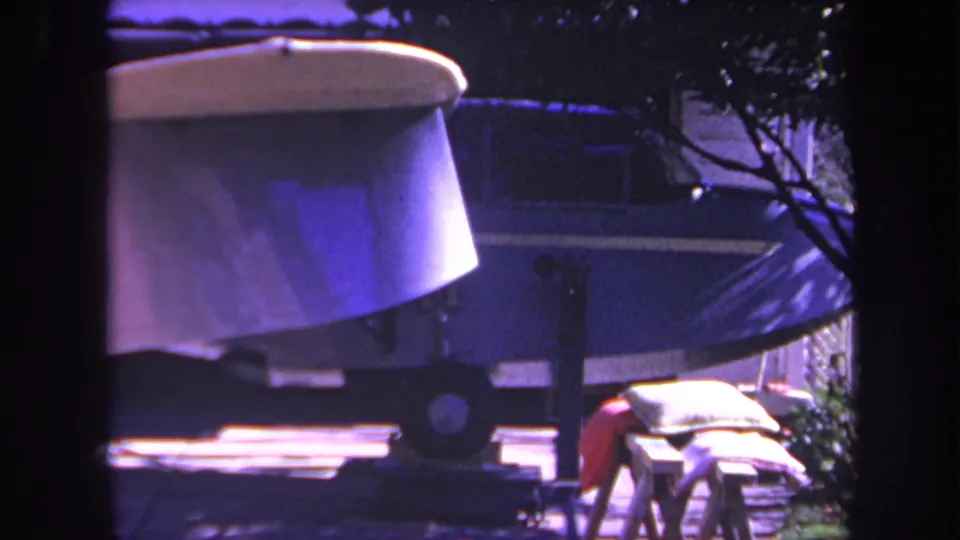 The frame shows the small blue aircraft in the same residential driveway view, with trailer wheels, a pale upper surface, nearby sawhorse, and dark vertical framing from the filming position.
