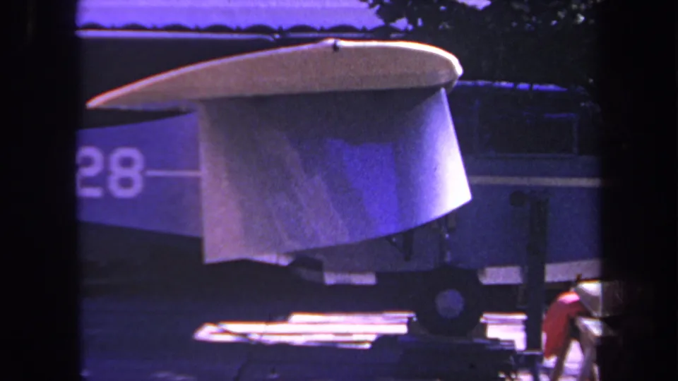 The final frame shows the small blue aircraft in the driveway with a partial number 28 visible on the side, a pale wing or tail surface above it, and dark framing at the sides of the shot.
