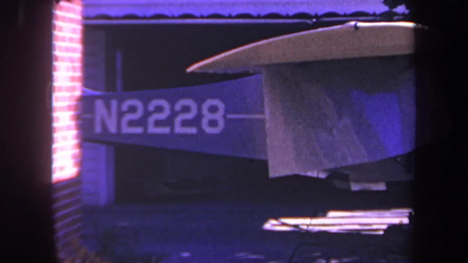 1969 Yucaipa home-movie frame with the plane tail, N2228 marking, red brick wall, and shadowed workshop area.
