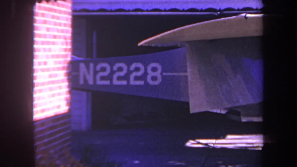 1969 Yucaipa frame showing the aircraft tail and N2228 marking beside brickwork, with strong purple-blue 8mm color shift.
