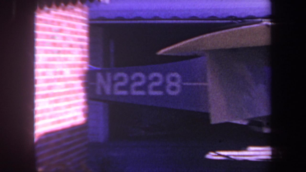 1969 8mm home-movie frame from Yucaipa, California showing the tail and wing of a small aircraft marked N2228 inside a garage or hangar-like structure.