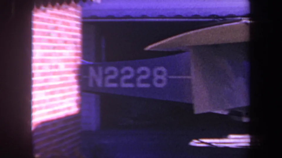 1969 8mm home-movie frame from Yucaipa, California showing the tail and wing of a small aircraft marked N2228 inside a garage or hangar-like structure.