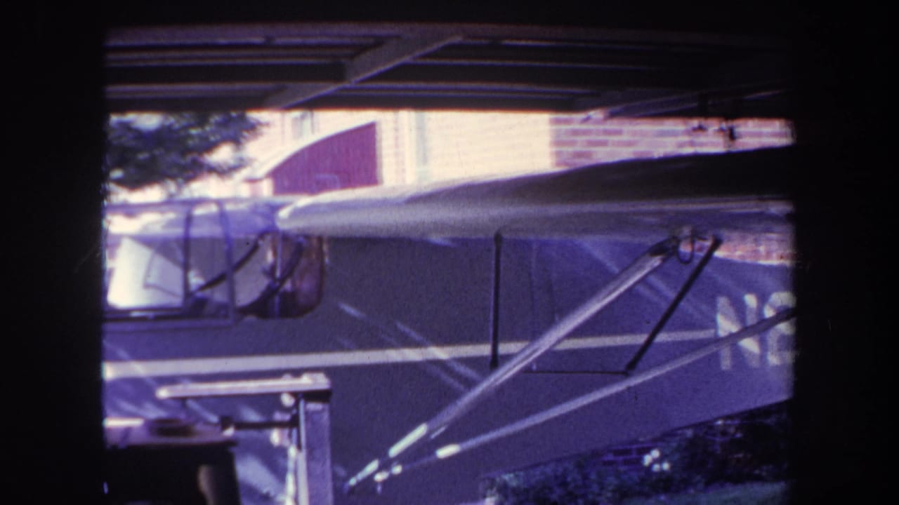 1969 Yucaipa 8mm frame showing a small aircraft under a carport, with cockpit windows, fuselage, wing struts, a brick wall, and tree foliage in view.