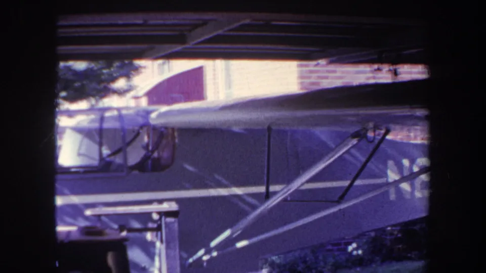 1969 Yucaipa 8mm frame showing a small aircraft under a carport, with cockpit windows, fuselage, wing struts, a brick wall, and tree foliage in view.