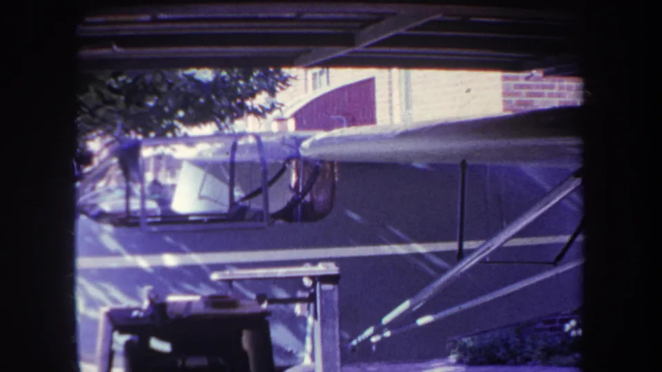 1969 Yucaipa home-movie frame with a small single-engine aircraft parked beneath a carport, showing the cockpit, fuselage side, wing edge, struts, and brick building behind it.