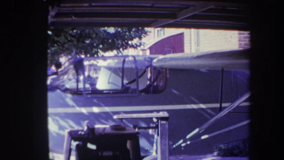 1969 Yucaipa 8mm frame showing the parked aircraft from a similar side angle, with cockpit glass, fuselage, wing struts, carport roof lines, and a brick wall visible.
