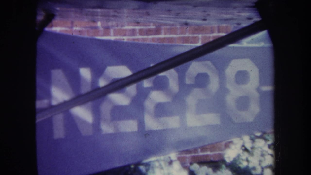 1969 8mm frame from Volmer Yucaipa, California, showing a blue aircraft tail or body panel with white N2228 lettering, partly obscured by dark aircraft structure and a brick wall behind it.