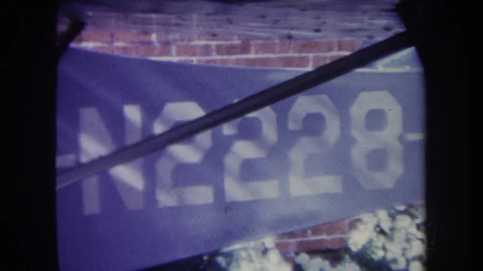 1969 8mm frame from Volmer Yucaipa, California, showing a blue aircraft tail or body panel with white N2228 lettering, partly obscured by dark aircraft structure and a brick wall behind it.