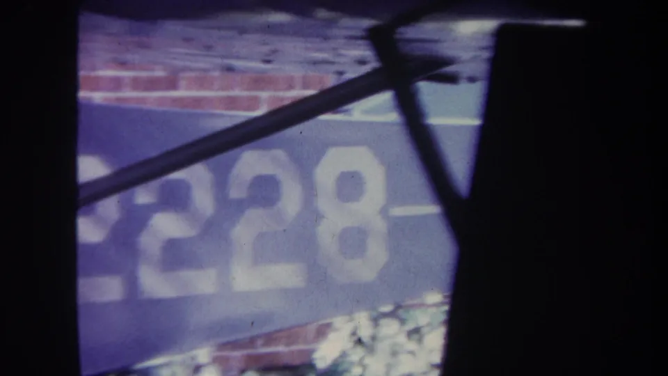 1969 8mm frame showing a cropped blue aircraft panel with partial white lettering, seen behind a dark vertical obstruction and angled aircraft structure near a brick wall.