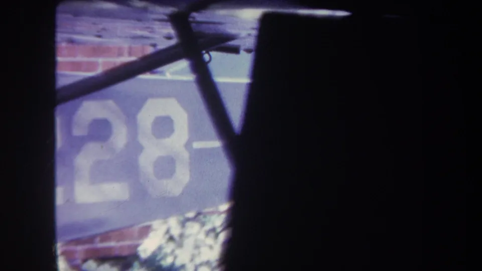 Final 1969 Volmer Yucaipa frame with part of the white aircraft marking visible on a blue panel, partly blocked by dark wing or fuselage shapes and set against a brick wall.