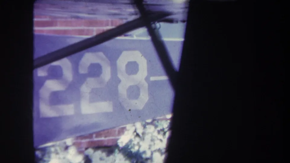 1969 8mm frame from Volmer Yucaipa showing a blue banner with white 228 lettering, a brick wall, dark foreground edges, and a diagonal metal bar.