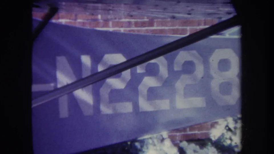 1969 8mm frame from Volmer Yucaipa showing white N2228 lettering on a blue banner, with brickwork above and dark foreground obstruction.