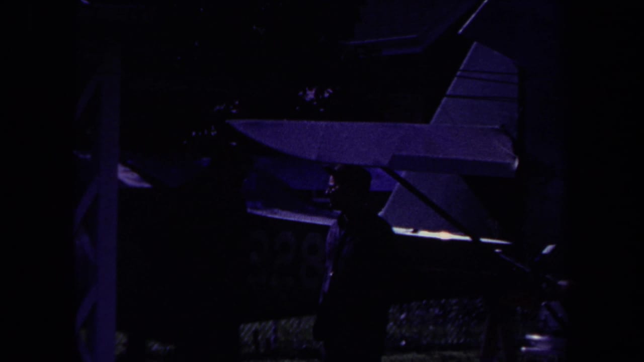1969 Yucaipa home-movie frame showing a dark-suited man near a small airplane under low purple light.