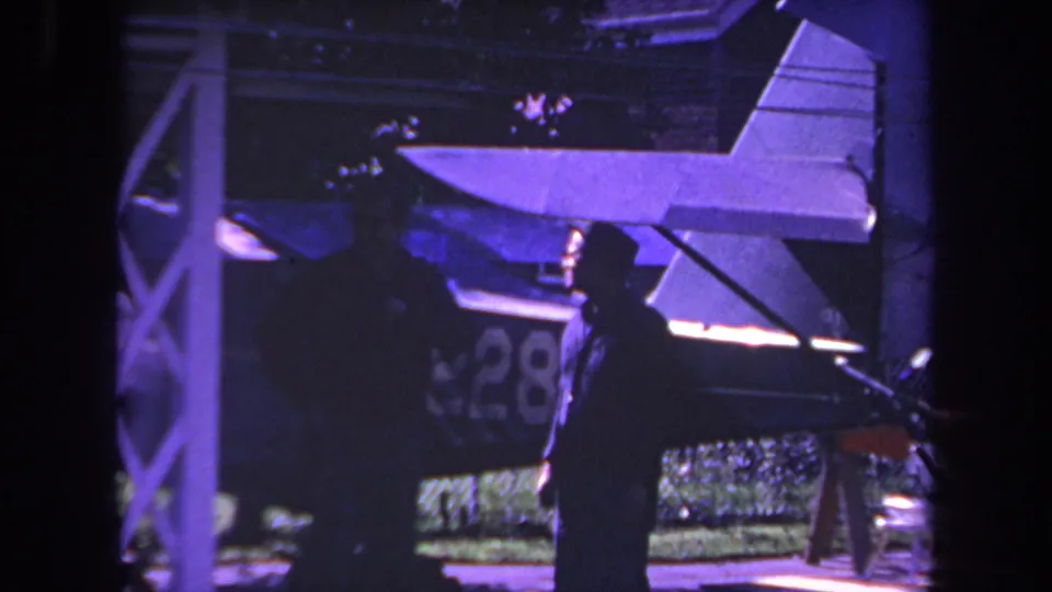 1969 Yucaipa home-movie frame with a man in dark clothing foregrounded against a small aircraft and foliage.