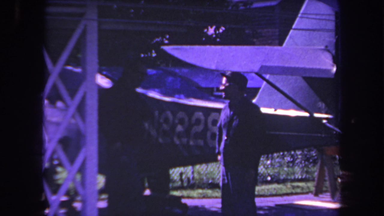 1969 Yucaipa home-movie frame showing two men in silhouette near a small aircraft under a runway-side structure.