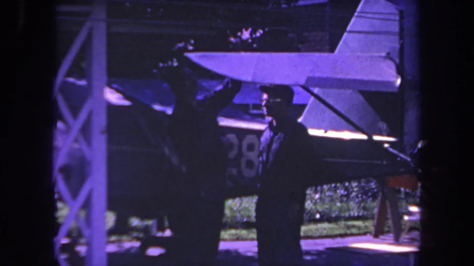 1969 Yucaipa-area 8mm frame showing two men near a small aircraft, with the plane tail, fuselage number, shelter post, and greenery visible.