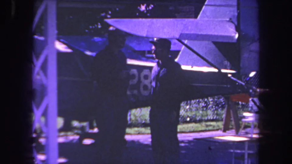 Purple-tinted 1969 8mm frame from the Yucaipa area showing two men beside a small plane, with a shelter post and the aircraft number partly visible.