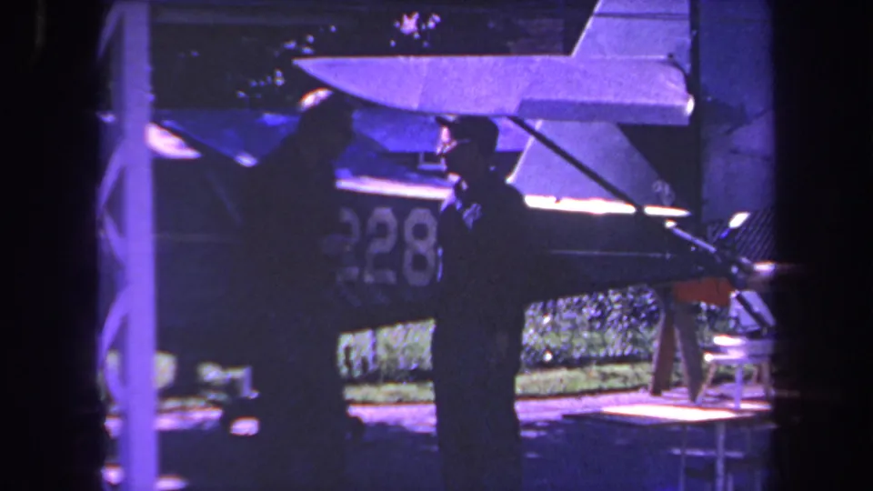 Final 1969 Yucaipa-area frame shows two men near a small aircraft under shade, with the tail, wing, fuselage number, and greenery behind them.