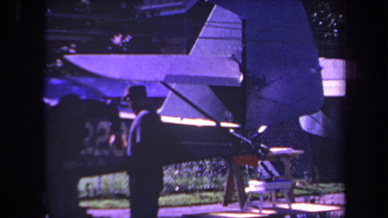 Two silhouetted men stand beside a small propeller plane under purple-blue light.