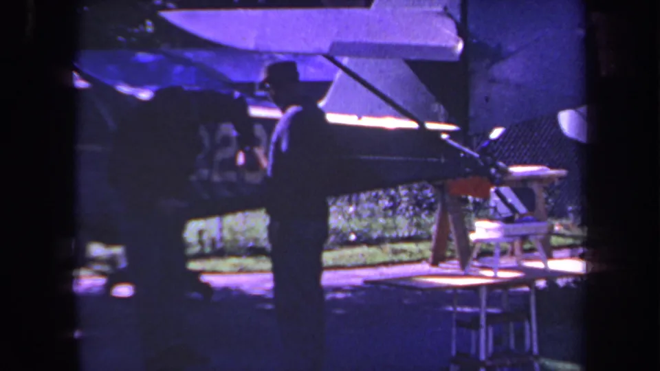1969 Yucaipa home-movie frame with a silhouetted man by a small propeller plane, aircraft surfaces, fence, and outdoor objects visible.