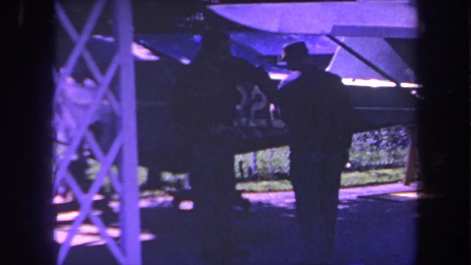1969 Yucaipa 8mm frame showing two men in silhouette near the side of a small propeller plane, with fence and yard details behind them.