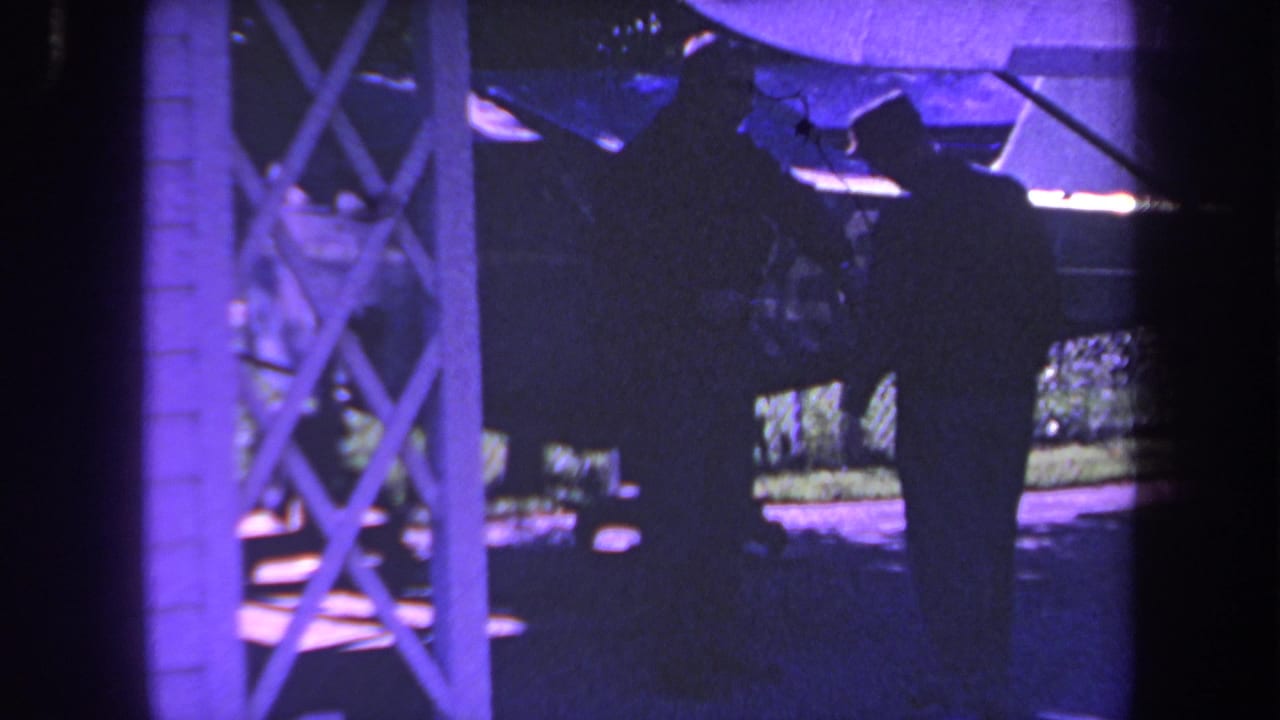 Two men in shadow stand beside a small aircraft under a covered structure.