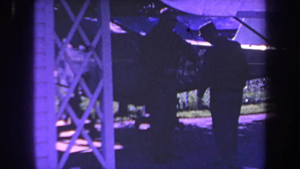 Two adult men in dark clothing stand near a small private aircraft under a covered outdoor structure in 1969 Yucaipa-area home-movie footage.