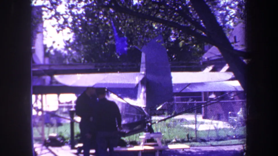 Two men appear beside the wing and tail of a small private aircraft, with trees and a chain-link fence visible in the background.
