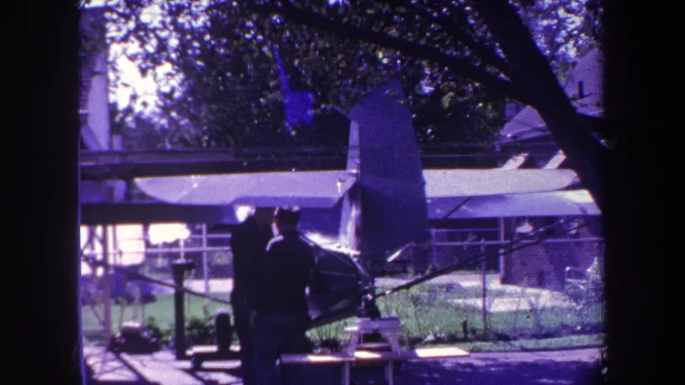 In the Yucaipa-area home-movie scene, two men face each other near the small aircraft, with the fence, grass, and trees behind them.