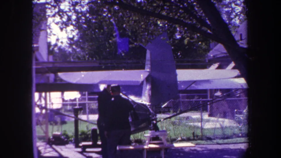 Two men remain near the small aircraft in the 1969 California home-movie footage, partly silhouetted against grass, trees, and a chain-link fence.