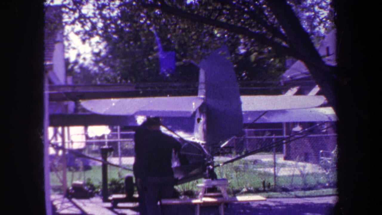 Man standing beside a small model airplane in a fenced backyard.