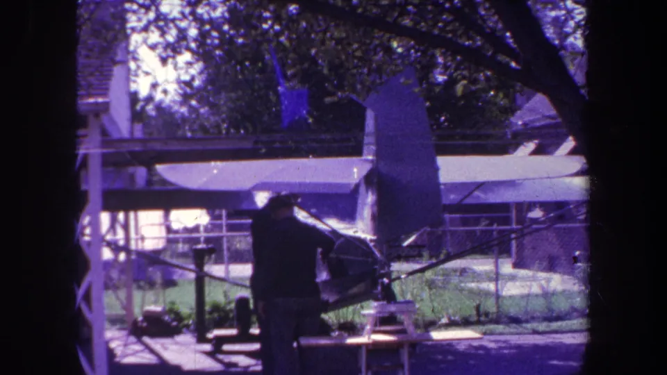 A man appears to adjust or inspect a model airplane in a Yucaipa backyard, partly framed by a dark tree trunk.