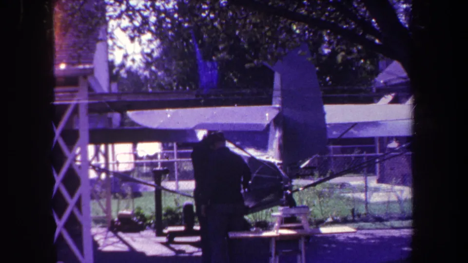 The home-movie frame shows the man and model airplane from a slightly shifted angle, with fence, grass, and house behind them.