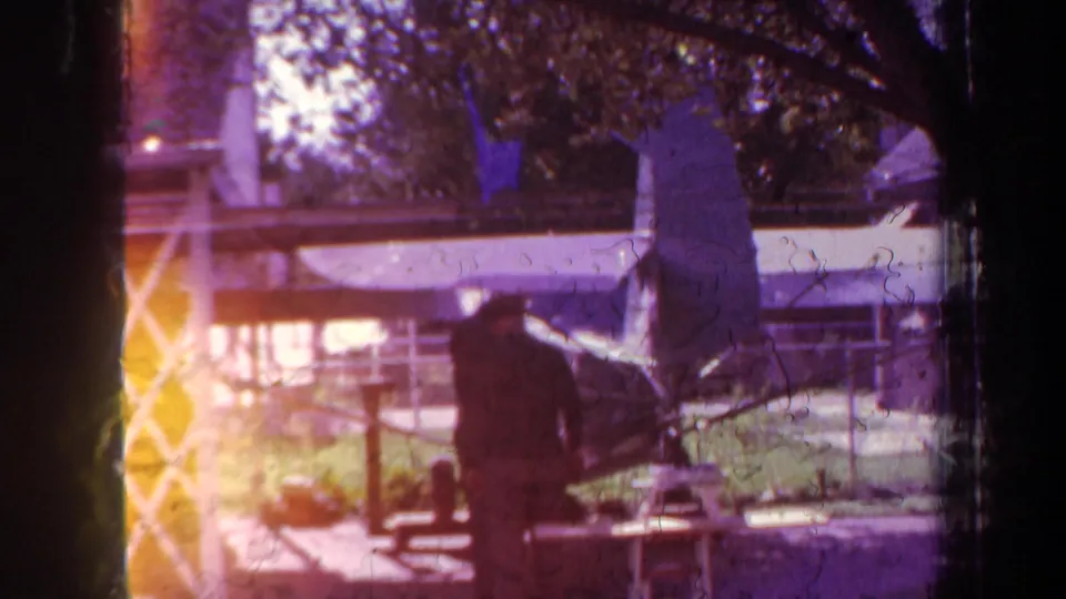 A color-shifted 1969 home-movie frame shows the man near the model airplane, with visible light flare and film wear.