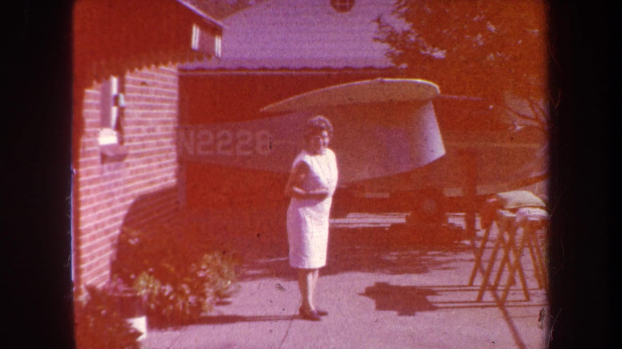 1969 Yucaipa home-movie frame showing a woman in a white dress on a driveway near a brick house and parked small aircraft.