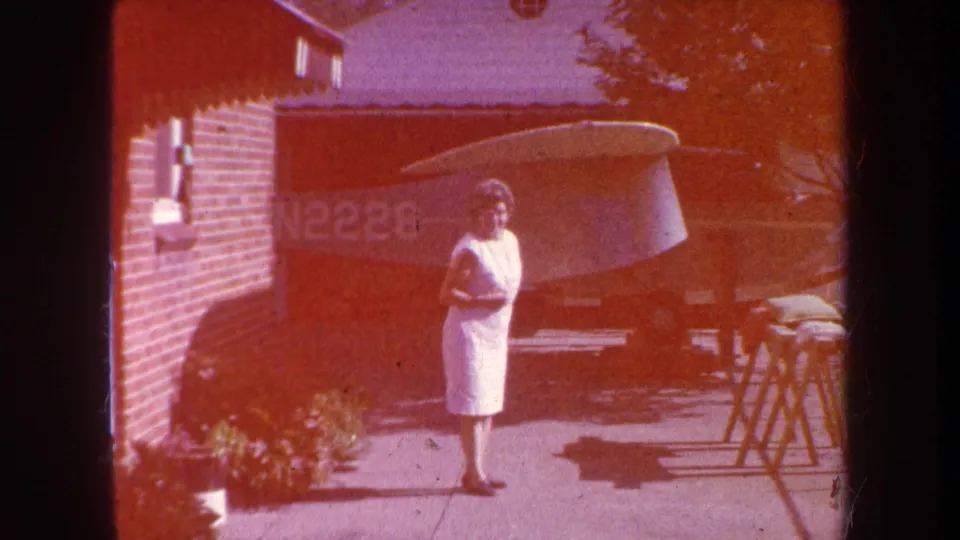 1969 Yucaipa home-movie frame showing a woman in a white dress on a driveway near a brick house and parked small aircraft.