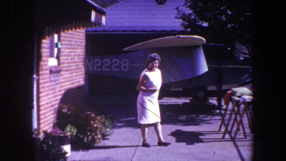 1969 Yucaipa home-movie still showing the woman moving across the driveway with the small aircraft behind her in shade.