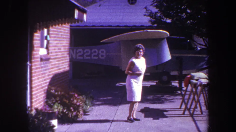 1969 Yucaipa frame of a woman near a residential driveway, looking toward the camera with a small aircraft parked behind her.