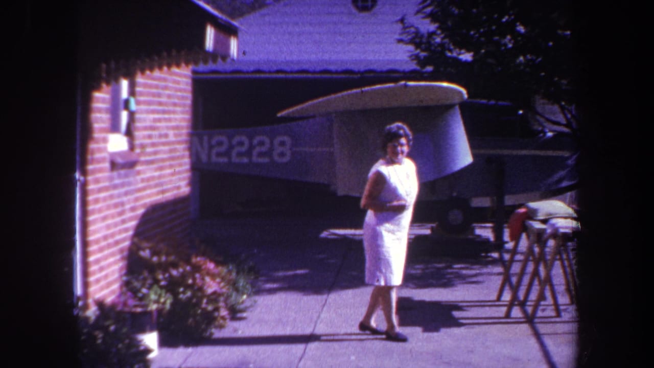 Woman in a white sleeveless dress walking on a sidewalk beside a brick house and parked airplane.